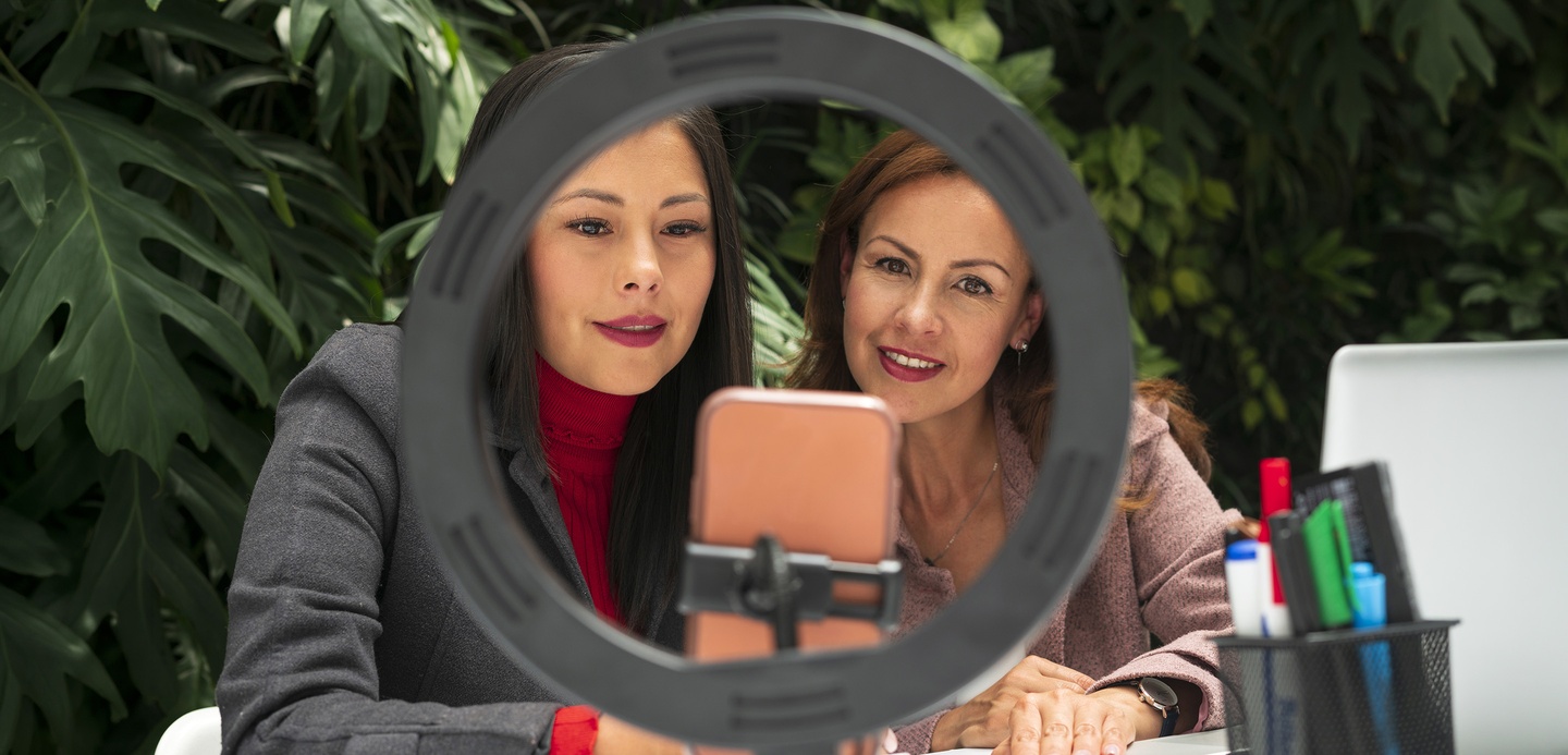 Two women sat at a desk, framed by a ring light, looking at a smartphone recording content. Two women sat at a desk, framed by a ring light, looking at a smartphone recording content.