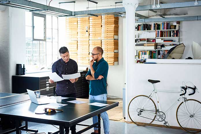 Two people standing in front of a ping pong table covered in paper, one person is holding two sheets of paper which both are looking at pensively Two people standing in front of a ping pong table covered in paper, one person is holding two sheets of paper which both are looking at pensively