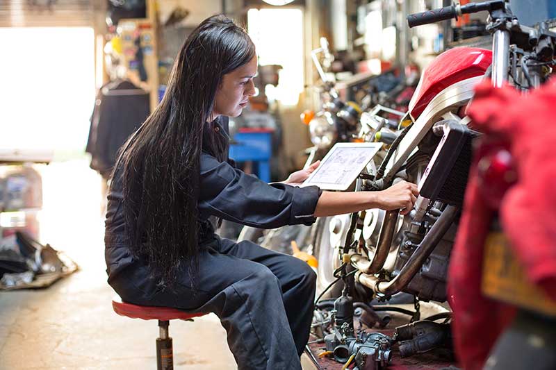 A person sitting on a stool wearing overalls holding a tablet and touching a motorbike in a garage A person sitting on a stool wearing overalls holding a tablet and touching a motorbike in a garage